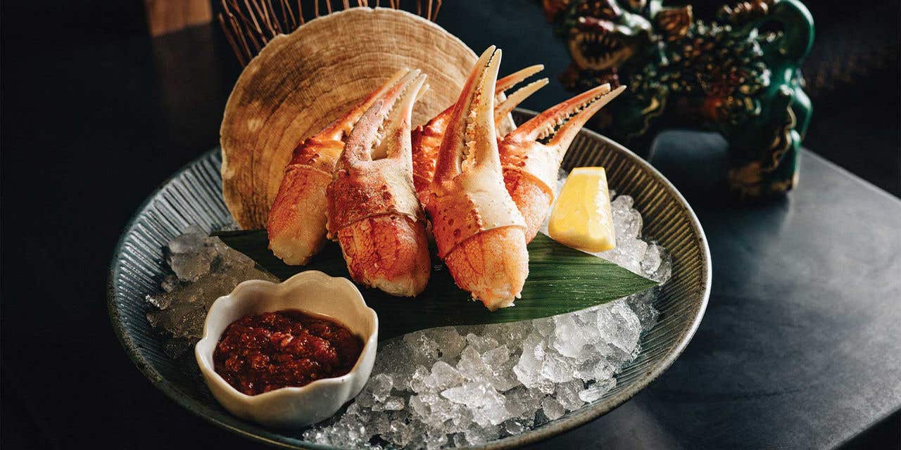 A group of crab claws over ice in a bowl with a side of red sauce and a lemon wedge.  