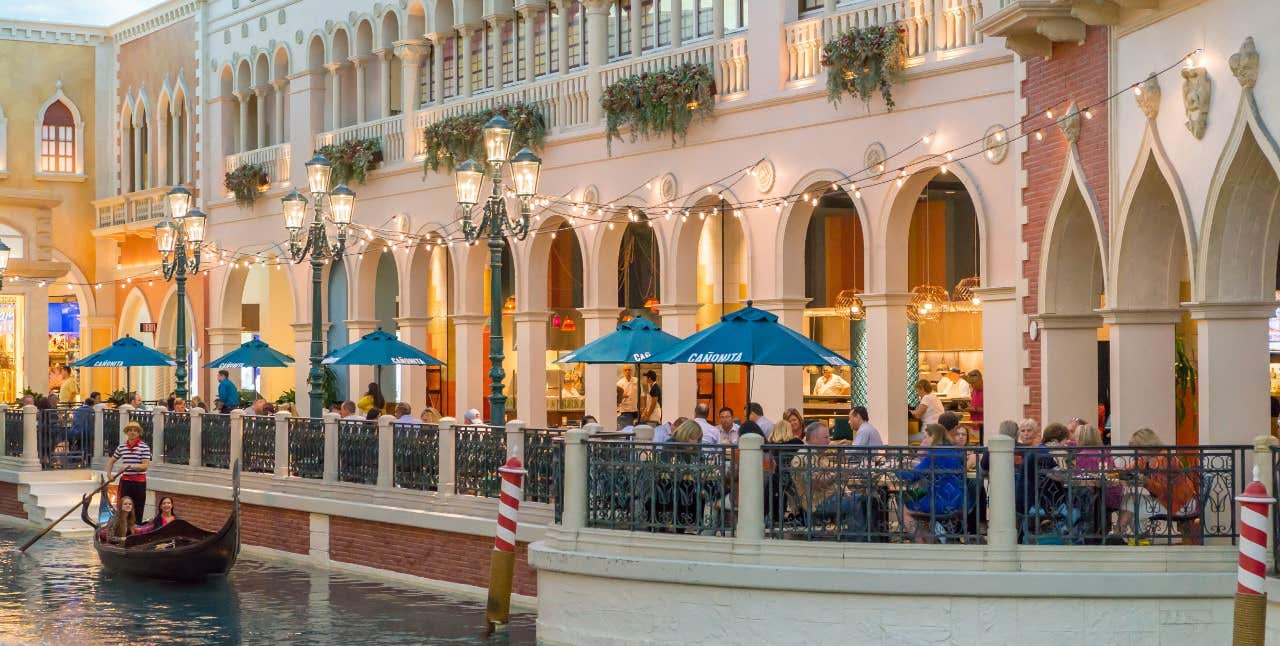 People enjoying dining outside of a Mexican restaurant next to a canal with a gondola in a plaza designed to resemble Venice.