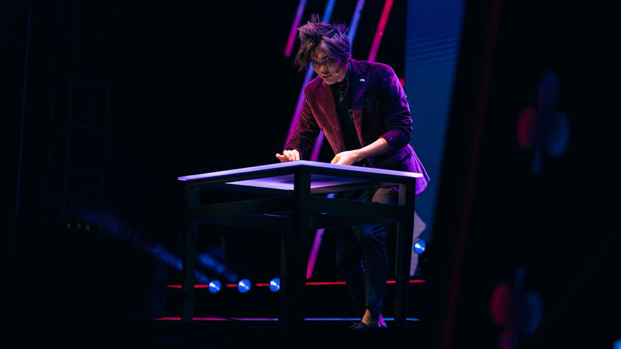 Shin Lim in a velvet jacket leaning over a table in a dimly lit, dramatic setting with colorful lighting.